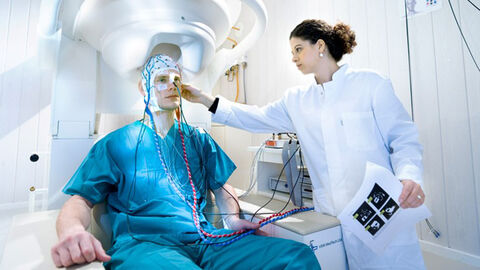 Wie funktioniert das Gehirn und wie lassen sich dessen Erkrankungen besser heilen? Ein Mann im blauen Hygieneanzug trägt eine Kappe, die Sensoren an seinem Kopf hält. Er sitzt unter einer Maschine mit einer Aussparung für seinen Kopf. Neben ihm steht eine Frau im weißen Kittel, die seine Maske justiert